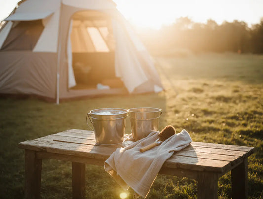 Camping gear and cleaning tools on a wooden table in warm sunlight after cooking outdoors, with a brush, towel, and bucket of soapy water beside a tent — calm scene showing the ritual of cleaning up after grilling.